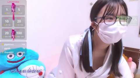 Media: Video of a young Asian woman with glasses, wearing a white surgical mask, and a white lab coat, kneeling in a brightly lit room. She has a serious expression.