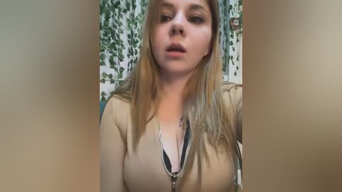 Media: A video of a young Caucasian woman with fair skin, long blonde hair, and a light brown jacket, standing indoors with a green leaf-patterned wall in the background.