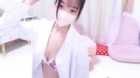 Media: A video of an East Asian woman with light skin, dark hair, wearing a white lab coat over a pink lace bra, a surgical mask, and polka-dotted pants, standing in a minimalist, brightly lit room with white walls and a bed.