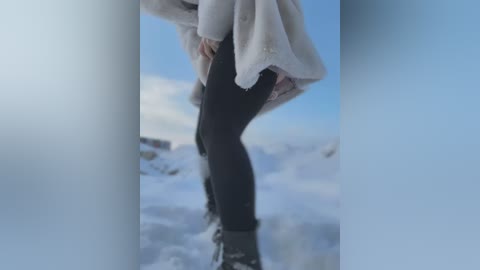 Media: Video of a person in black leggings, white sweater, and black boots, standing in a snowy outdoor setting with a clear blue sky. The image is focused on the lower half of the subject's body.