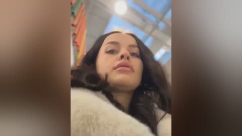 Media: A video of a young woman with long dark hair, fair skin, and full lips, wearing a beige sweater, standing in an industrial space with orange and white walls and large windows.