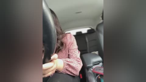 Media: Video of a woman with long brown hair, wearing a pink patterned sweater, driving a car. Her left hand grips the steering wheel, while the car's interior features black seats and a beige dashboard.