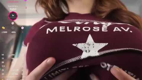 A video of a woman lifting her maroon shirt, revealing her ample breasts, which are covered in white stars. The background is blurry, possibly an indoor setting with a teal wall.