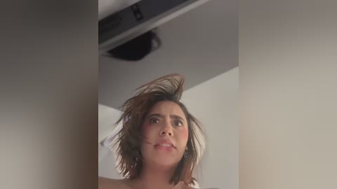 Video of a woman with medium brown hair, slightly tousled, looking upward with a surprised expression. She has a light tan complexion and is wearing a white top. Background shows a plain, off-white wall and a ceiling vent.