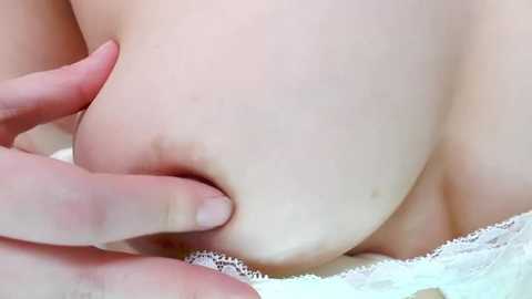 Media: A close-up video of a Caucasian woman's pale, smooth skin, showing a mole on her breast. Her hand gently squeezes the breast, revealing a white lace bra. The background is out of focus, emphasizing the texture and color contrast.