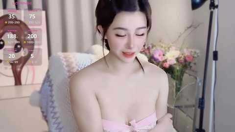 Media: Video of an Asian woman with fair skin, dark hair in a bun, wearing a pink strapless dress, sitting on a chair with a floral cushion, surrounded by a room with a colorful bouquet and a digital clock.