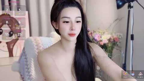 Media: Video of a young Asian woman with long black hair, fair skin, and a slender physique, sitting in a cozy room with a floral arrangement, a lamp, and a calendar in the background.