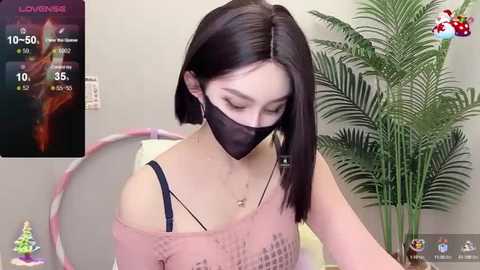 Media: A video of a young Asian woman with straight black hair, wearing a black face mask and a pink off-shoulder top, sitting in front of a green potted plant, with a digital thermometer showing 10.5\u00b0C on the left.