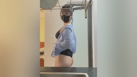 Media: A video of a woman with medium skin tone, wearing a blue shirt, black bra, and black panties, using a pull-up bar in a gym, with a yellow wall and equipment in the background.