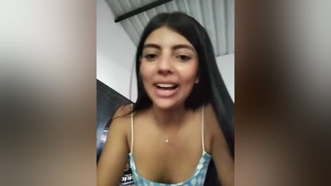 Media: Video of a young Latina woman with long black hair, wearing a low-cut blue and white patterned camisole, smiling and laughing, indoors with corrugated metal ceiling and white walls.