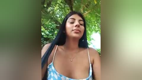 Media: Video of a Latina woman with long black hair, medium skin tone, wearing a blue and white polka-dot spaghetti-strap top, standing in a lush green forest, looking serene.