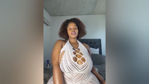 Media: Video of a plus-sized Black woman with natural hair, wearing a white, intricate, halter-neck dress, sitting on a bed in a modern, minimalist bedroom.