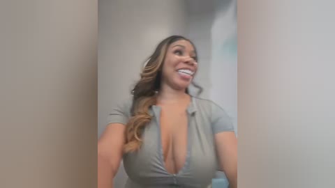 Media: Video of a smiling, light-skinned woman with long, wavy brown hair, wearing a low-cut gray shirt that reveals cleavage, standing in a narrow hallway.