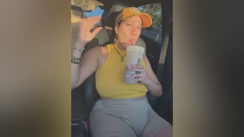 Media: A video of a curvy, light-skinned woman with a yellow tank top and grey shorts, wearing a yellow cap and a necklace, sipping a drink while sitting in a car.