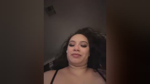 Media: A video of a plus-sized, light-skinned woman with curly hair, wearing a black bra, making a pouty face. The background is dimly lit with a ceiling light and shadowy wall.