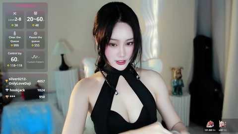 Media: Video of an Asian woman with long black hair, wearing a revealing black halter top, standing in a dimly-lit bedroom with a bed and a lamp.