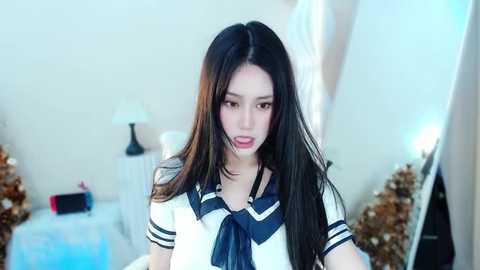 Media: Video of an Asian woman with long black hair, wearing a schoolgirl outfit, in a softly lit bedroom with Christmas decorations.