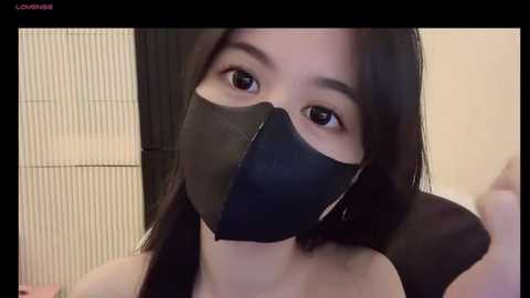 Media: Video of an East Asian woman with long black hair, wearing a black face mask, sitting against a beige wall with a black partition in the background.