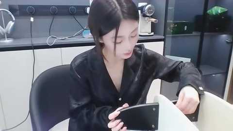 Media: Video of an Asian woman with straight black hair, wearing a black leather jacket, intently looking at a black smartphone on a white table in a modern, minimalist kitchen with white cabinets and black countertops.