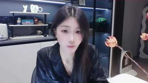 Media: Video of an Asian woman with long black hair and fair skin, wearing a black leather jacket, sitting in a modern kitchen with sleek black cabinets and white appliances.