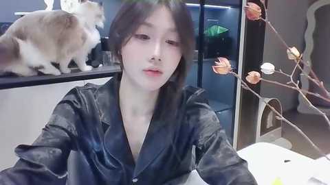 Media: Video of a young East Asian woman with fair skin, dark hair, and a slim build, wearing a black leather jacket. She sits in a modern room with a beige cat on a shelf, a floral-patterned curtain, and a white table.