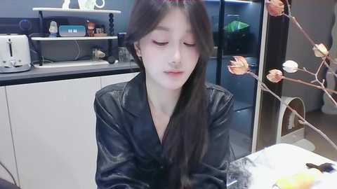 Media: Video of a young Asian woman with long black hair, wearing a black leather jacket, sitting at a table in a modern kitchen with white cabinets, a black countertop, and decorative branches.