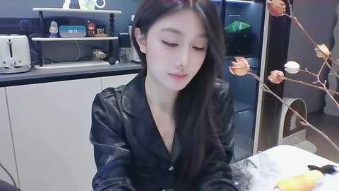 Media: Video of a young East Asian woman with long black hair, wearing a black leather jacket, sitting at a white table in a modern kitchen. The background features a black countertop with appliances and decorative items.