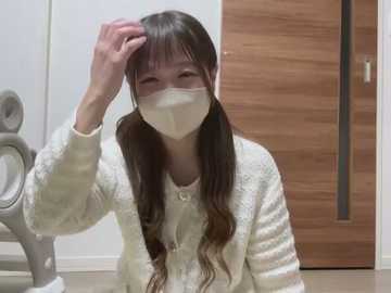 Media: Video of an East Asian woman with long dark hair, wearing a white mask, white sweater, and sitting on a tiled floor. Background includes wooden door and gray washing machine.