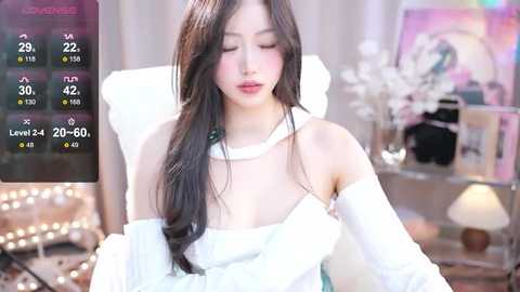 Media: Video of a young Asian woman with long black hair, wearing a white off-shoulder dress, sitting indoors with a white cushion and floral decor in the background.