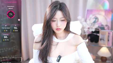 Media: Video of an Asian woman with long, straight black hair, wearing a white off-shoulder top, looking down, in a brightly lit room with pastel decor.