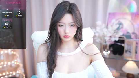 Media: Video of a young East Asian woman with long, straight black hair, wearing a white off-shoulder sweater, sitting indoors. Background features a blurry, softly lit room with white curtains, a lamp, and decorative items.