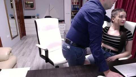 Video of a man in a navy blue shirt leaning over a woman in a black striped top and jeans, seated at a desk in an office with white chairs, shelves, and a window.