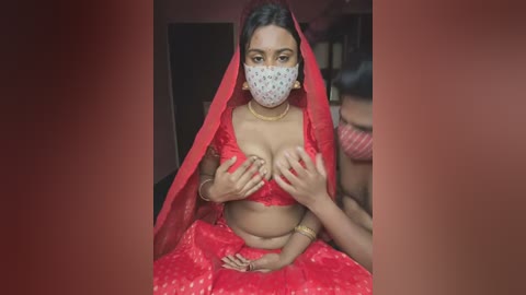 Media: Video of a South Asian woman in a red sari, covering her breasts with her hands, wearing a white face mask, set against a blurred background.