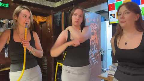 Media: Video of three women in a bar: two blonde with short hair, one brunette with long hair. All wear black tank tops and beige pants, using a yellow hose, and standing near a graffiti-covered wall.