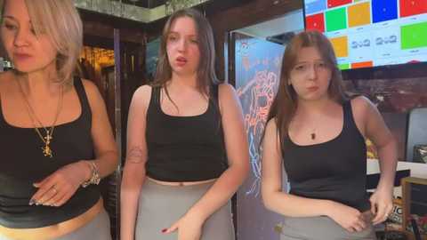 Media: Video of three young women in black tank tops and high-waisted shorts, one with blonde hair, one with brunette hair, and one with auburn hair, in a lively, colorful bar.