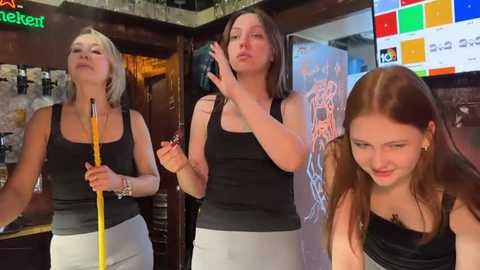 Media: Video of three women in casual black tank tops and white pants, dancing in a dimly-lit bar with colorful neon lights, one holding a yellow baton.