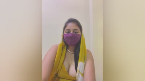 Media: A video of a middle-aged woman with a medium skin tone, wearing a yellow saree, purple face mask, and a silver necklace, standing in a narrow, beige-walled corridor.