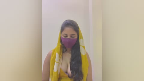 Media: A video of a woman with long black hair, wearing a yellow and white sari, purple mask, and necklace, standing against a plain, light-colored wall.