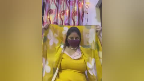 Media: Video of a woman with medium skin tone, wearing a yellow saree with white floral patterns, a purple face mask, and a light pink blouse. She holds the saree, revealing a colorful curtain with red and purple hues in the background.