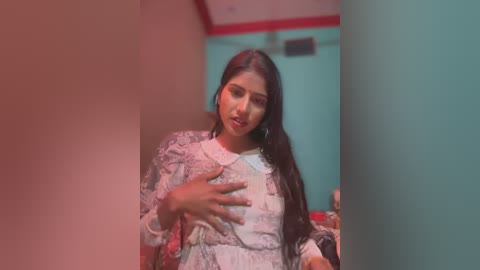 Media: Video of a young South Asian woman with long black hair, wearing a floral-patterned saree, posing in a dimly lit room with teal walls and red trim.