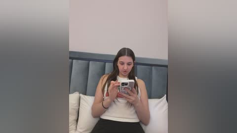 Media: Video of a young woman with long brown hair, wearing a white tank top and black skirt, sitting on a bed with a grey headboard, holding a phone, looking at it with a neutral expression.