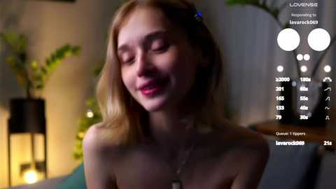 Media: A video of a smiling, topless blonde woman with fair skin, wearing a blue hair accessory, in a dimly lit room with a potted plant and a black lamp.