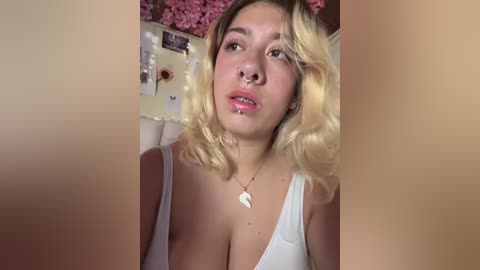 Media: Video of a young woman with wavy blonde hair, fair skin, and a nose ring, wearing a low-cut white top, in a cozy room with pink flowers and fairy lights on the wall.