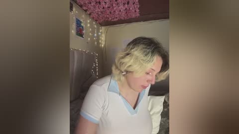 Media: Video of a blonde woman with short, wavy hair, wearing a white polo shirt with blue trim, standing in a dimly lit bedroom adorned with fairy lights, pink floral wallpaper, and a tufted headboard.