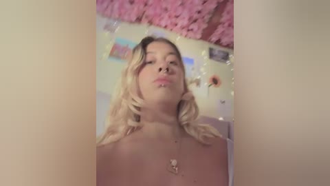 Media: Video of a topless, fair-skinned woman with blonde hair, wearing a nose ring, and making a pouty face. Background features pink cherry blossom garlands and colorful posters.