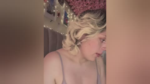 Media: A video of a young woman with light skin and blonde hair, wearing a light gray tank top, captured from the side, against a pink wall with fairy lights and framed pictures.