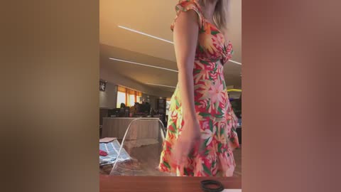 Media: Video of a woman in a colorful floral dress standing in a bright, modern kitchen with beige walls, stainless steel appliances, and a glass-top table.