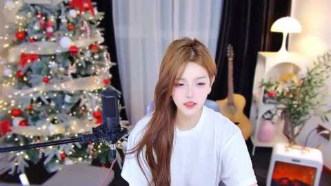 Video of a young woman with long, wavy brown hair, wearing a white T-shirt, standing indoors near a decorated Christmas tree and a guitar.