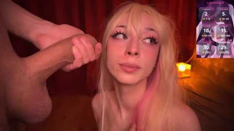 Video of a pale-skinned, blonde-haired young woman with pink streaks, wearing a top, kneeling, gazing up at an erect, circumcised penis. Background includes a dimly lit, red-curtained room with a lit candle.