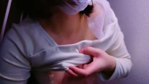 Media: Video of a woman lifting her white shirt, revealing a pink bra and cleavage, with a white headband, in a dimly lit room.